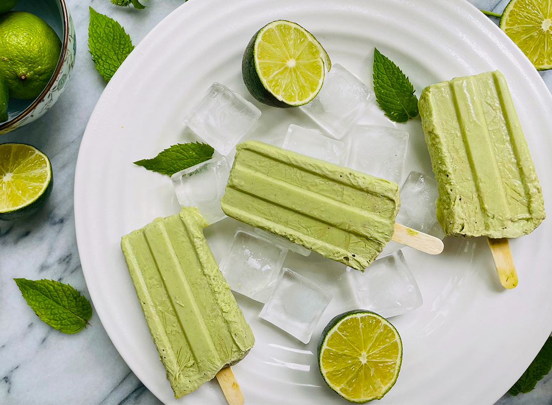 3 green Key Lime Coconut Popsicles on a plate with ice, mint leaves and fresh lime